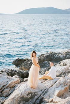 Bride stands half-turned on the rocky seashore near the seated groom. High quality photo