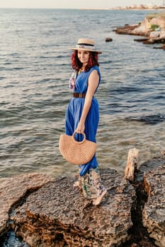 A woman in a dress, hat and with a straw bag is standing on the beach enjoying the sea. Happy summer holidays.