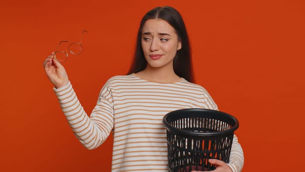 Lovely woman taking off, throwing out glasses into trash bin after medical vision laser treatment therapy surgery, looking smiling at camera. Young adult girl isolated alone on red studio background