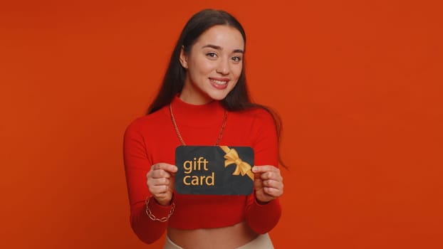Young woman in crop top showing pointing on gift discount certificate voucher coupon for store holidays sale. Lovely girl with shopping christmas surprise card isolated alone on red studio background