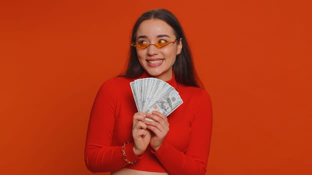 Rich pleased boss millennial woman waving money dollar cash banknotes bills like a fan, success business career, lottery winner, big income, wealth. Young pretty girl isolated on red background