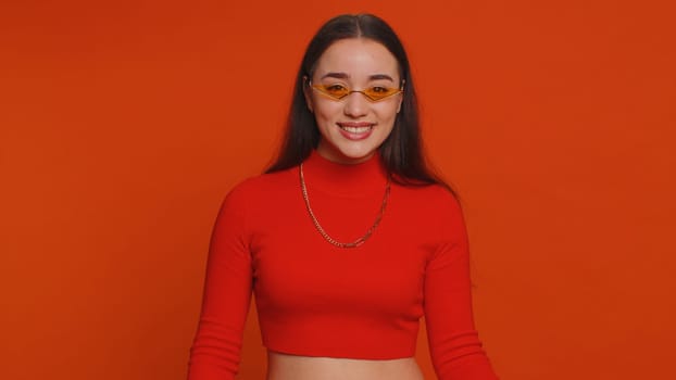 Portrait of happy pretty young woman in crop top wearing sunglasses, looking at camera with toothy charming smile, flirting, expressing optimism. Millennial girl isolated on red studio background