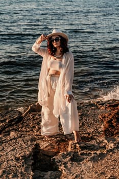 A woman in a white pantsuit and hat is standing on the beach enjoying the sea. Happy summer holidays.