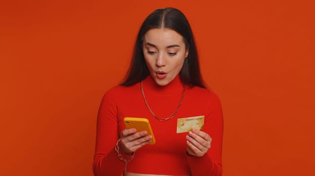 Sincere pretty woman customer using credit bank card and smartphone while transferring money, purchases online shopping, payment. Finance and internet. Young girl isolated alone on red background