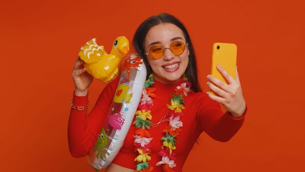 Happy young traveler blogger woman in modern sunglasses with swimming ring, luggage taking selfie on smartphone with subscribers record make video call online. Lovely tourist girl on red background