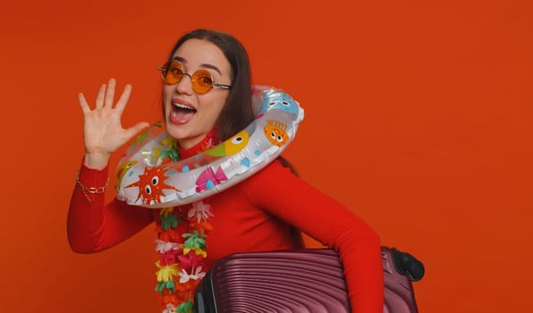 Happy curly haired woman in sunglasses, hawaiian lei walking along with luggage bag, swimming ring. Young smiling girl dancing, on red studio background. Summer vacation, journey, trip to seaside