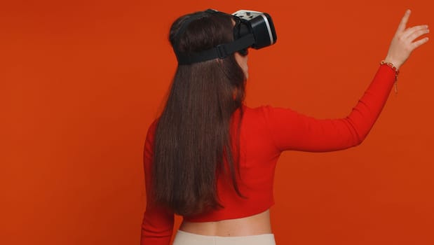 Woman using headset helmet to play simulation game app online. Watching virtual reality 3D 360 video content. Girl in VR goggles isolated on studio red background. Futuristic technology. Rear view