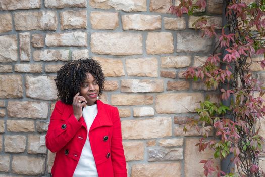 African american woman in red coat talking on smartphone near brick wall, technology use concept, wi-fi connection. High quality photo