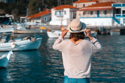 A young girl in a hat looks at the picturesque view with the sea and moored yachts on a sunny day, a backpacker travels and discoveries beautiful places on the coast during her holidays.