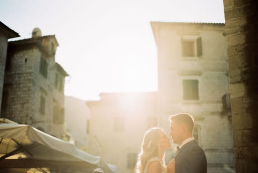 Groom almost kisses bride against the backdrop of old houses in bright sunlight. High quality photo
