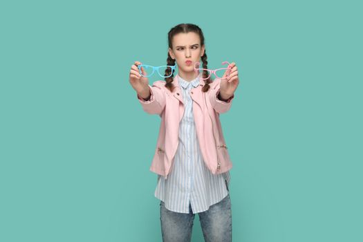 Portrait of funny crosseyed teenager girl with braids wearing pink jacket holding two eyewear, having very bad eyesight, choosing eyeglasses. Indoor studio shot isolated on green background.