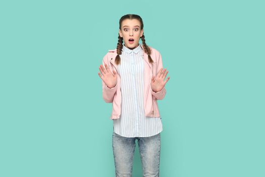 Portrait of scared young teenager girl with braids wearing pink jacket looking at camera with big eyes and open mouth, showing stop gesture. Indoor studio shot isolated on green background.