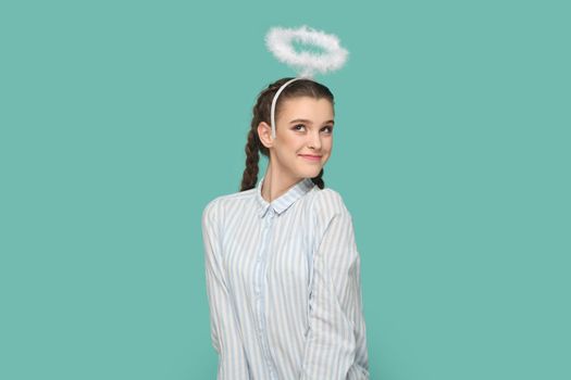 Portrait of pretty lovely friendly angelic teenager girl with braids wearing striped shirt and nimb over head, coquetry with boyfriend. Indoor studio shot isolated on green background.