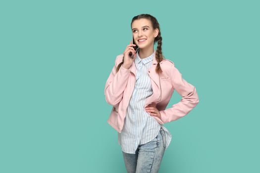 Portrait of positive smiling teenager girl with braids wearing pink jacket standing and talking on mobile phone, having pleasant conversation. Indoor studio shot isolated on green background.