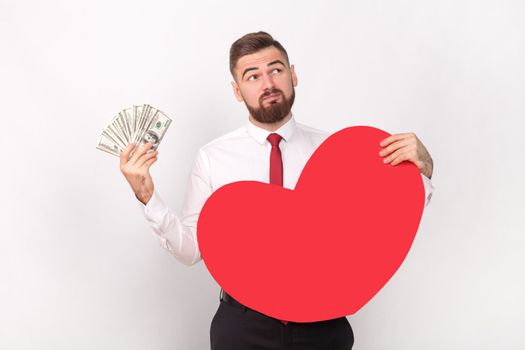 Portrait of puzzled confused pensive bearded man wearing white shirt and tie, holding big dollar banknotes and big red heart, looking away. Indoor shot isolated on gray background.