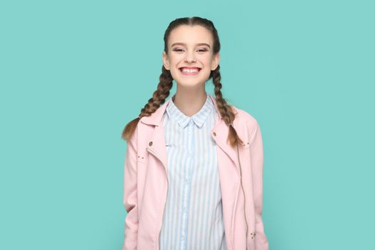 Portrait of extremely happy joyful teenager girl with braids wearing pink jacket standing looking at camera with toothy smile, smiles broadly. Indoor studio shot isolated on green background.