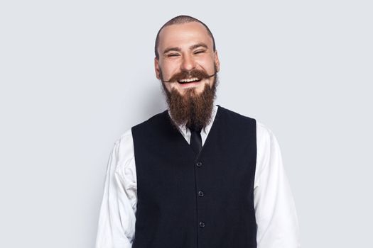 Portrait of excited attractive young adult man with beard and mustache standing and laughing, hearing funny jokes, looking at camera. Indoor studio shot isolated on gray background.