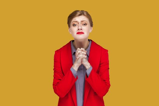 Portrait of hopeful woman with red lips standing with hands together, pleading, asking to forgive her, looking at camera, wearing red jacket. Indoor studio shot isolated on yellow background.