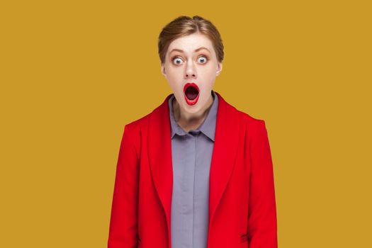 Portrait of shocked surprised astonished woman with red lips standing looking at camera with big eyes and open mouth, wearing red jacket. Indoor studio shot isolated on yellow background.