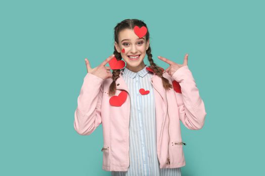 Portrait of romantic smiling teenager girl with braids wearing pink jacket standing pointing at little red heart on her body, expressing happiness. Indoor studio shot isolated on green background.