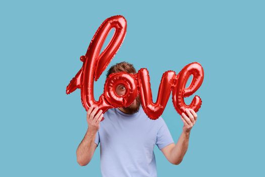 Portrait of romantic bearded man hiding his face behind love word of foil balloons, expressing his feelings, spying, being fall in love. Indoor studio shot isolated on blue background.