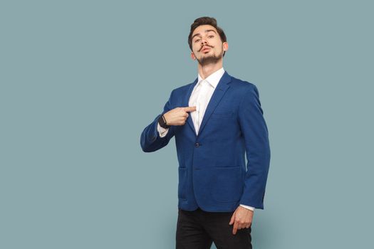 Portrait of self confident proud man with mustache standing pointing at himself, looking at camera with pride, wearing white shirt and jacket. Indoor studio shot isolated on light blue background.