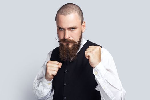 Portrait of serious angry man with beard and mustache standing with clenched fists, being ready to fight with somebody, looking at camera. Indoor studio shot isolated on gray background.
