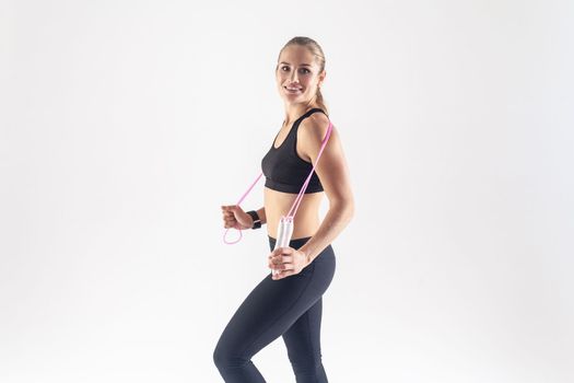 Side view portrait of happy athletic blonde woman holding skipping rope and smiling toothily at camera, wearing black fitness clothing. Indoor studio shot isolated on gray background.