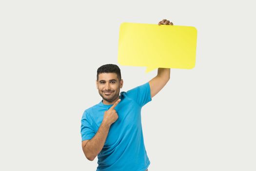 Portrait of optimistic smiling unshaven man wearing blue T- shirt standing pointing at yellow speech frame, space for words and thoughts. Indoor studio shot isolated on gray background.