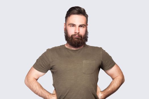 Portrait of bearded man wearing dark green T-shirt looking cross-eyed with stupid facial expression, fooling around, keeps hands on hips. Indoor studio shot isolated on light gray background.