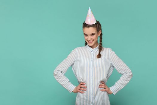 Portrait of happy festive teenager girl with braids wearing striped shirt and party cone, celebrating her birthday, keeps hands on hips. Indoor studio shot isolated on green background.