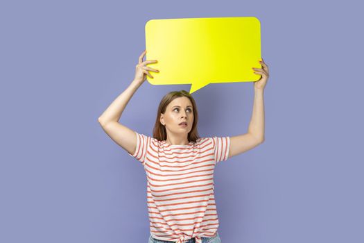Portrait of pensive blond woman wearing striped T-shirt looking away and holding yellow bubble thought over her head, copy space for text. Indoor studio shot isolated on purple background.