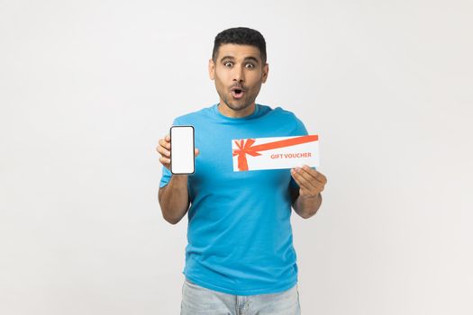 Portrait of shocked astonished man wearing T- shirt standing with gift voucher and showing smart phone with blank screen, copy space for advertisement. Indoor studio shot isolated on gray background.