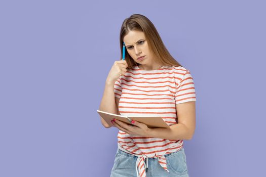 Portrait of pensive thoughtful blond woman wearing striped T-shirt writing in paper notebook, thinking about task, keeps pen near temple. Indoor studio shot isolated on purple background.