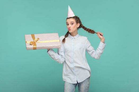 Flirting delighted attractive teenager girl with braids wearing striped shirt and party cone, holding her present on birthday, sending air kisses. Indoor studio shot isolated on green background.