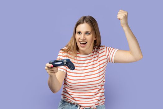 Portrait of excited amazed blond woman wearing striped T-shirt holding in hands gamepad joystick, clenched fist, showing yes gesture, winning. Indoor studio shot isolated on purple background.
