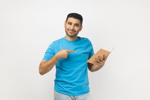 Portrait of attractive positive young adult unshaven man wearing blue T- shirt standing pointing index finger at interesting book. Indoor studio shot isolated on gray background.