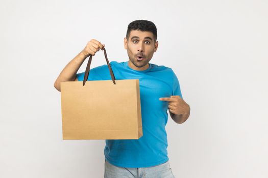 Portrait of shocked surprised astonished unshaven man wearing blue T- shirt standing pointing at shopping bag, great purchase in mall. Indoor studio shot isolated on gray background.