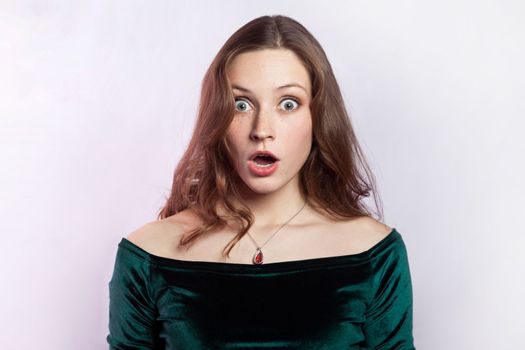 I can't believe this. Portrait of shocked astonished woman wearing green dress, looking at camera with big eyes and open mouth, being very surprised. Indoor studio shot isolated on gray background.