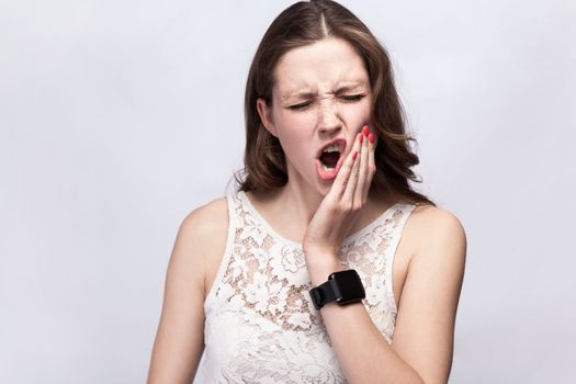 Portrait of stressed woman wearing white dress standing with frowning face and closed eyes, feeling terrible toothache, needs to visit dentist. Indoor studio shot isolated on gray background.