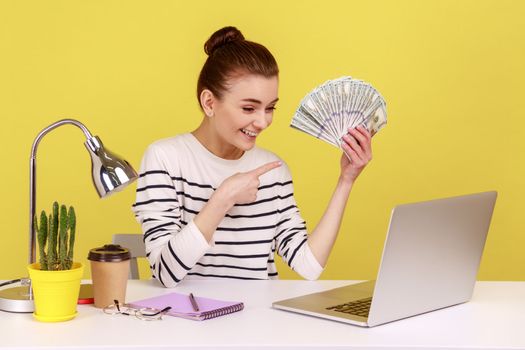Positive young woman pointing finger at hundred dollar bills, holding sitting at workplace, looking at laptop, high salary, bonuses and perks. Indoor studio studio shot isolated on yellow background.