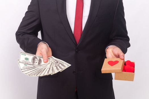Portrait of unknown anonymous man wearing black official style suit, white shirt and red tie, holding little box with hearts and fan of dollar banknotes. Indoor studio shot isolated on gray background
