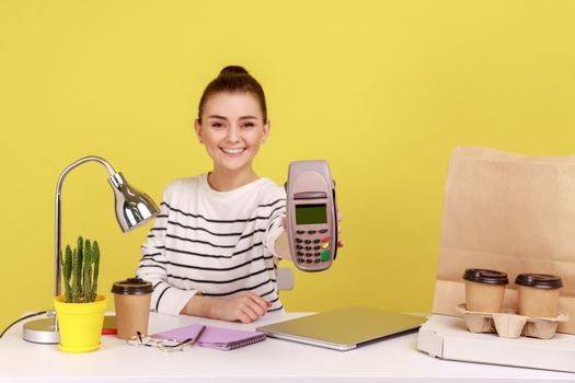 Smiling positive woman holding in hand pos contactless payment terminal, using paypass for purchases payment while sitting on workplace. Indoor studio studio shot isolated on yellow background.