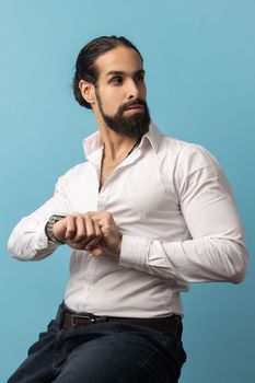 Handsome confident man with beard wearing white shirt sitting and touching his wristwatch, looking away, being harry to important event. Indoor studio shot isolated on blue background.