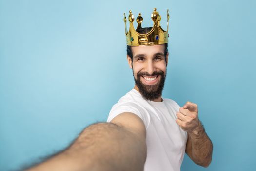 Portrait of smiling man with beard wearing white T-shirt and gold crown, pointing at camera, POV, point of view of photo, expressing happiness. Indoor studio shot isolated on blue background.