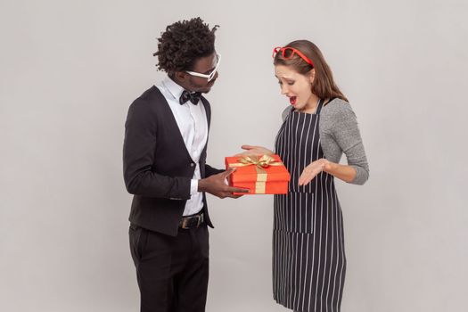 Portrait of woman and man in white glasses celebrating anniversary, husband giving to amazed surprised wife red present box. Indoor studio shot isolated on gray background.