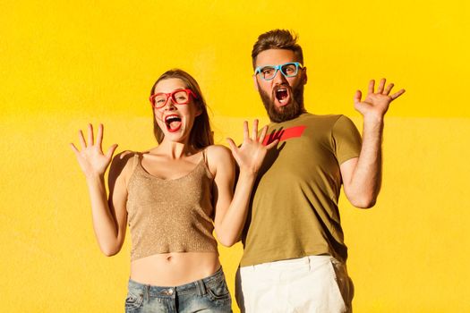 Portrait of funny young adult man and woman standing with raised arms and scared facial expression, being frighten, looking at camera. Indoor studio shot isolated on yellow background.