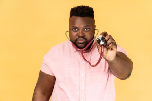 Portrait of serious bearded man wearing pink shirt standing looking at camera, holding stethoscope, listening lungs or hear beating. Indoor studio shot isolated on yellow background.