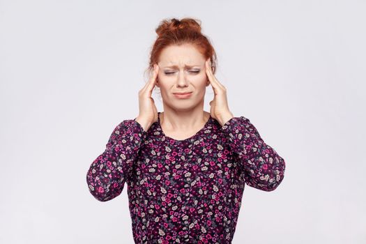 Portrait of unhealthy sick young adult redhead woman wearing dress massaging temples, suffering from headache, migraine, feels unwell. Indoor studio shot isolated on gray background.