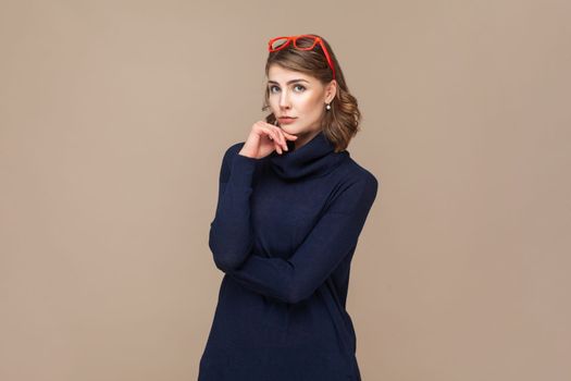 Portrait of pensive thoughtful woman with wavy hair in red red glasses on her head holding her chin, thinking about future. Indoor studio shot isolated on light brown background.